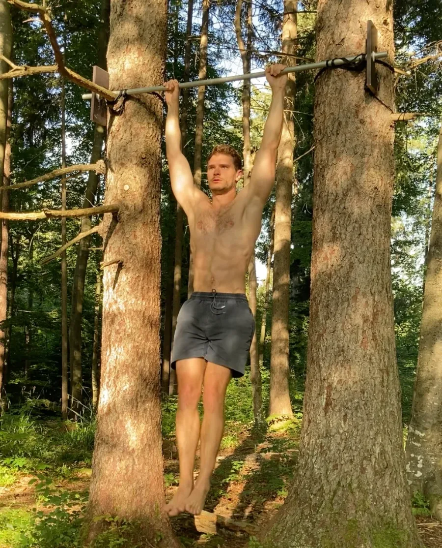 Nils Enders-Brenner mid pull-up on a bar rigged between two trees in the forest at golden hour