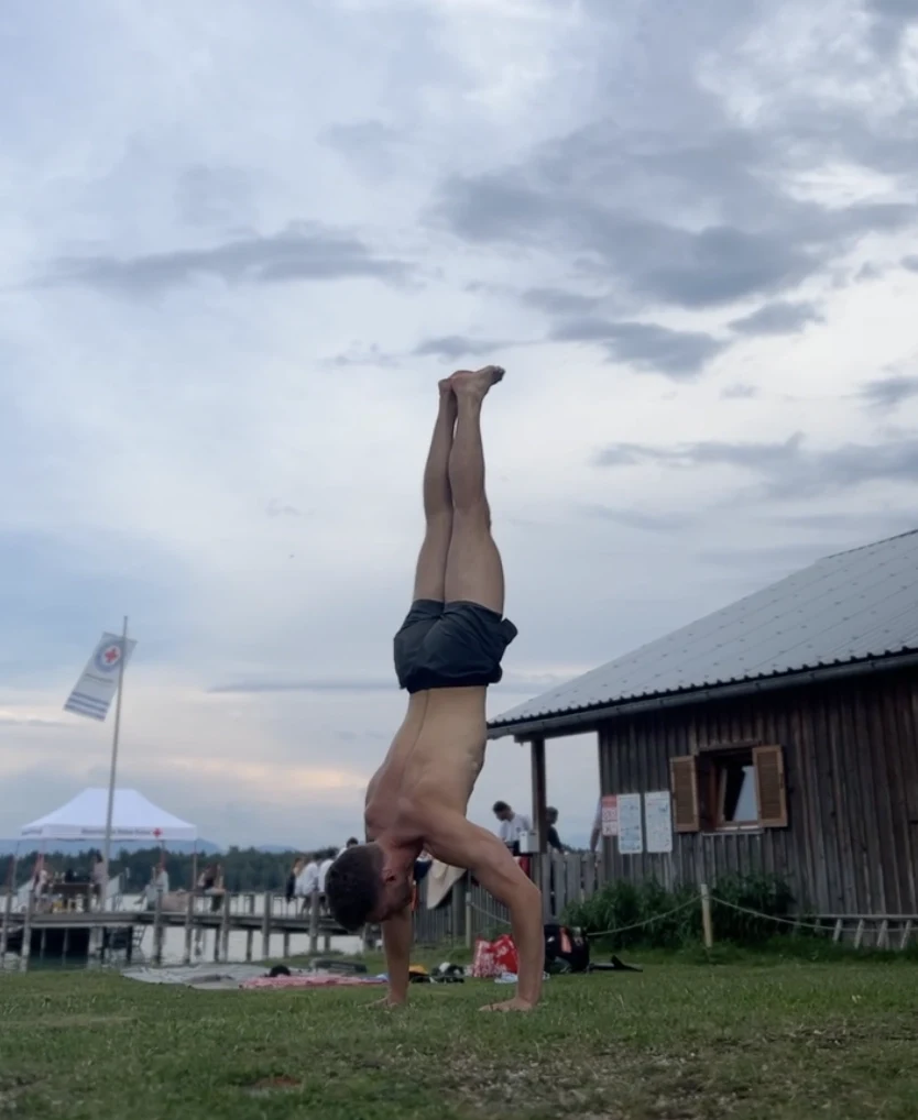 Nils Enders-Brenner holding a handstand on grass, a lake and boathouse in the background